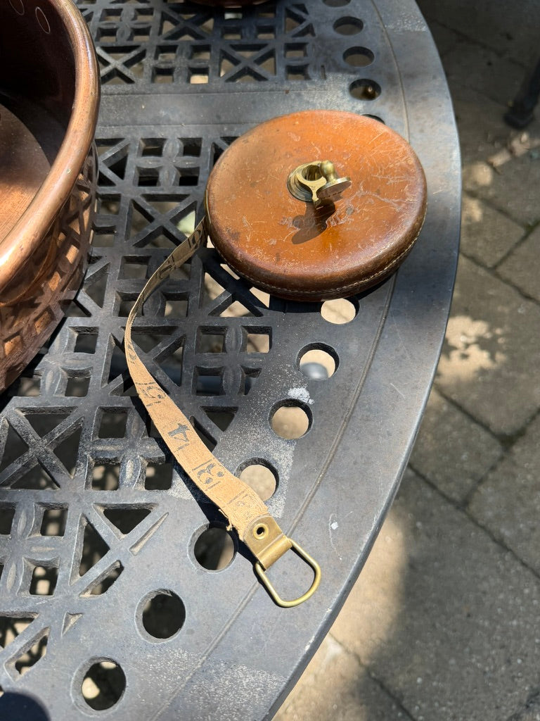 A vintage copper measuring tape reel with a brass hook and a tan measuring strap, resting on a black metal patio table outdoors.