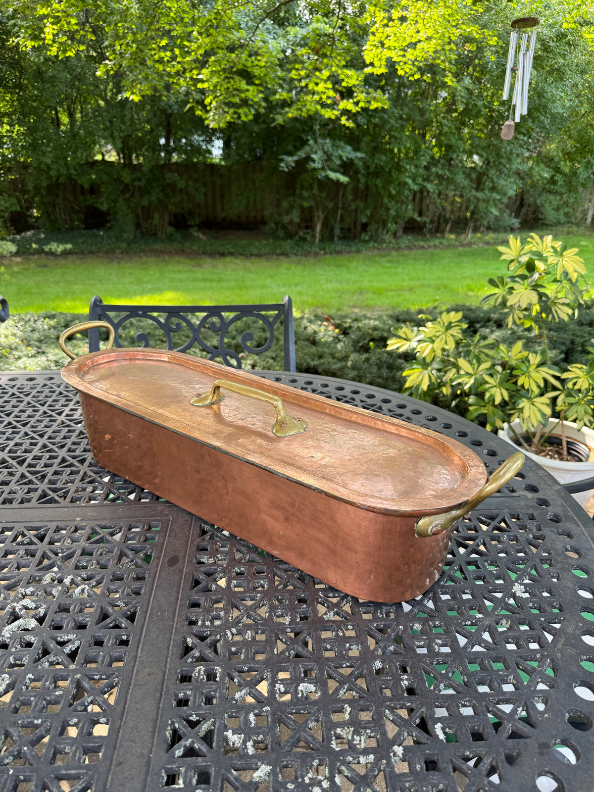 A vintage copper lidded tub with brass handles sits on a black metal patio table in a backyard garden setting.