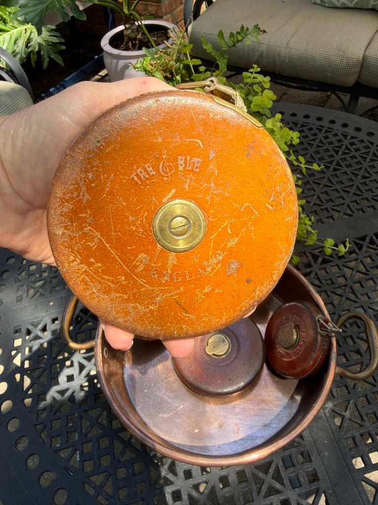 A vintage leather circular case with a brass button and embossed text, tape measure tool 