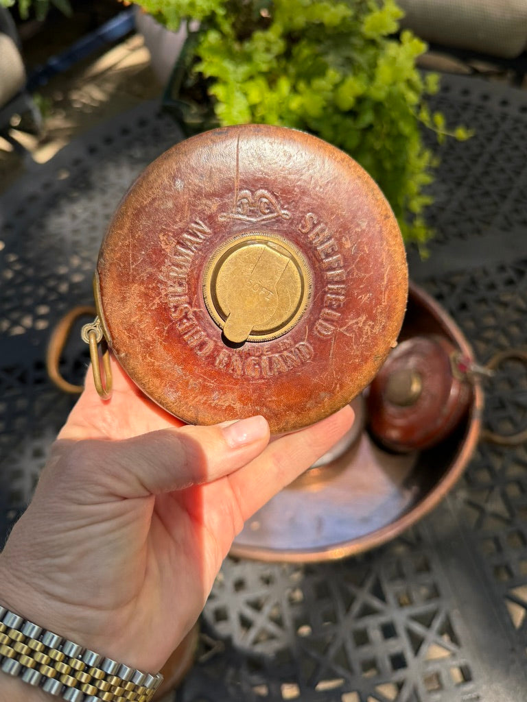 A hand holding a round, weathered copper-colored metal tin with embossed text reading 'SHEFFIELD ENGLAND' and a brass-colored center emblem, set against an outdoor patio background with a metal table and greenery.