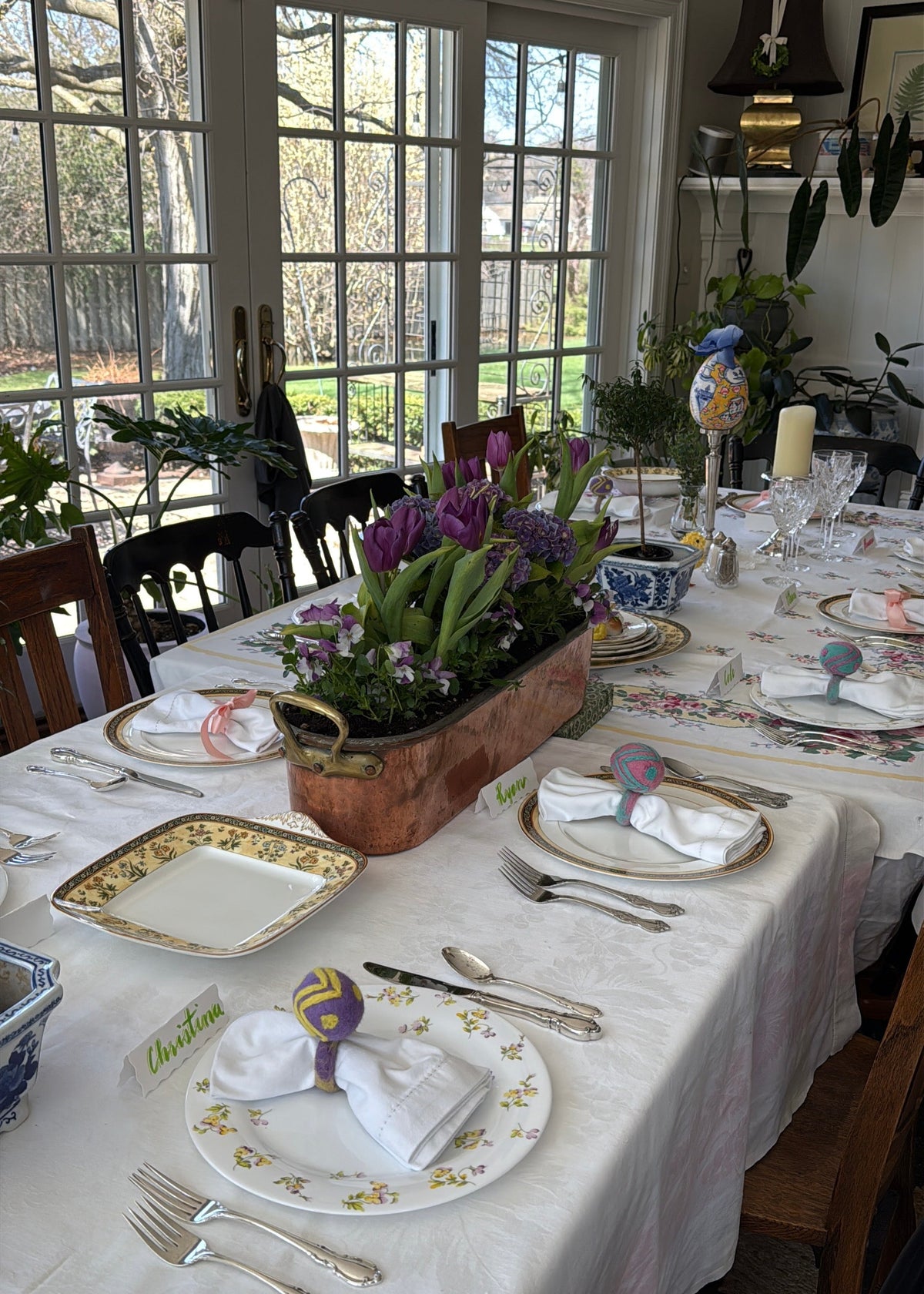 A dining table set for a meal with a central copper fish poacher planter filled with purple flowers and green leaves.