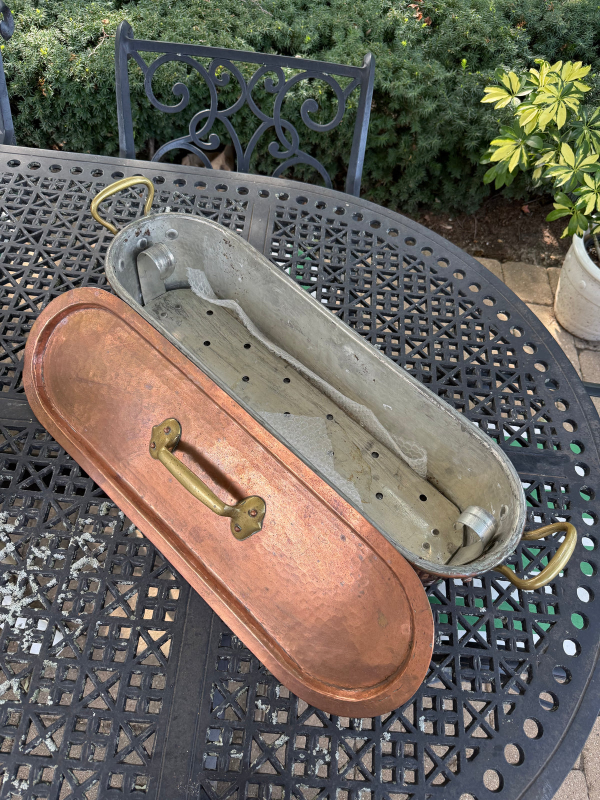 A vintage copper lidded container with a brass handle, shown next to an open weathered gray metal tub with brass handles, placed on a black metal patio table outdoors.