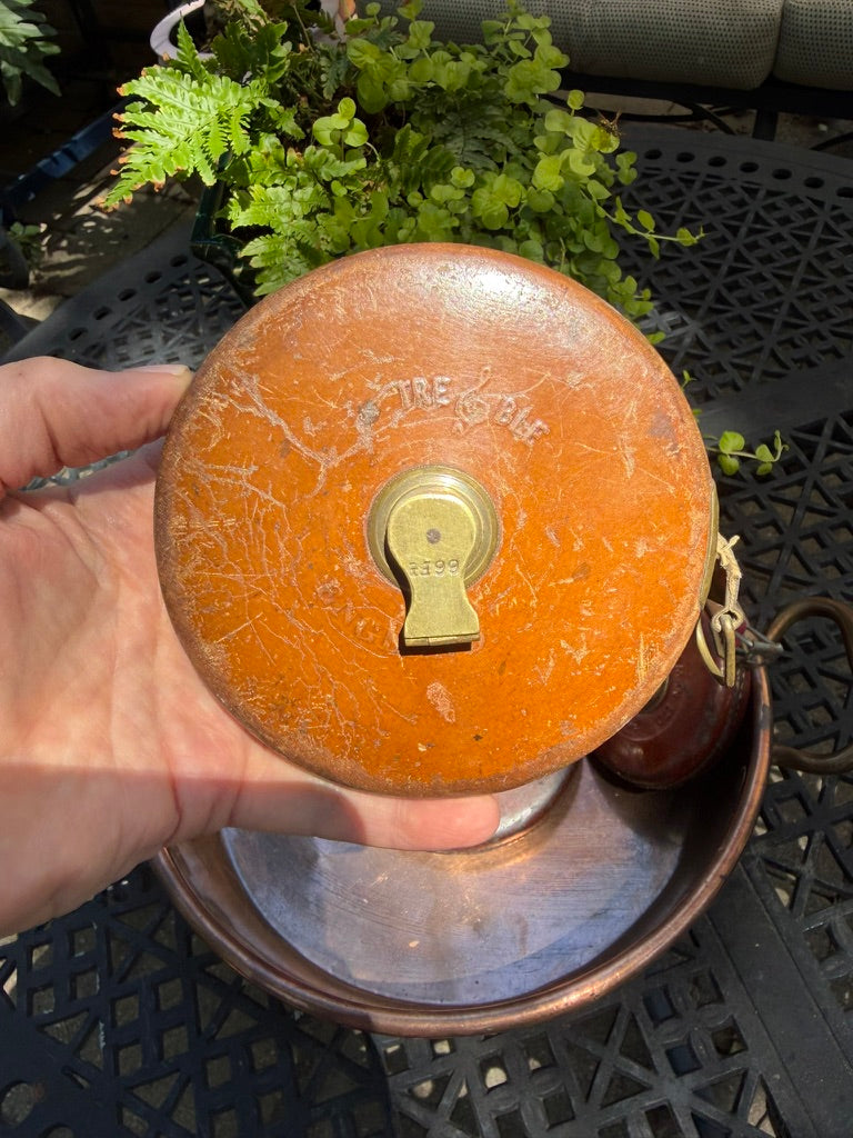 A hand holding a vintage orange fire hydrant cover with a brass keyhole detail, set against an outdoor patio background with green plants and a black metal table.