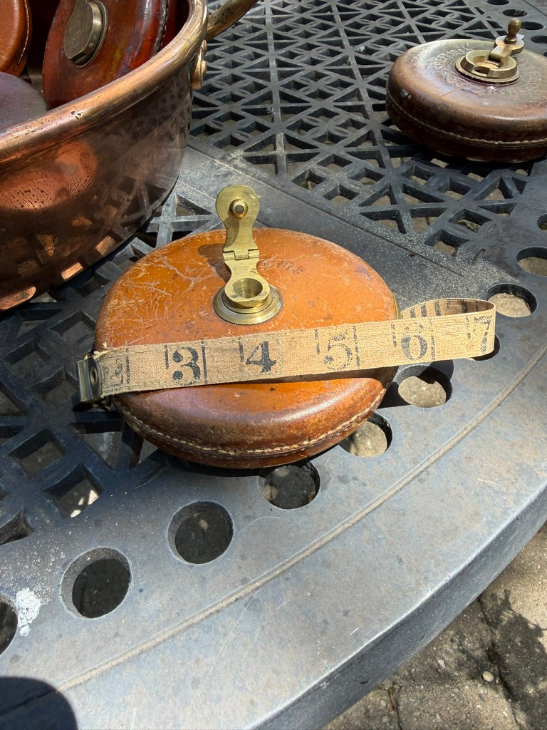 A vintage copper and leather measuring tool with a brass plunger and a beige tape measure wrapped around it, resting on a black metal lattice table.