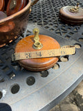 A vintage copper and leather measuring tool with a brass plunger and a beige tape measure wrapped around it, resting on a black metal lattice table.
