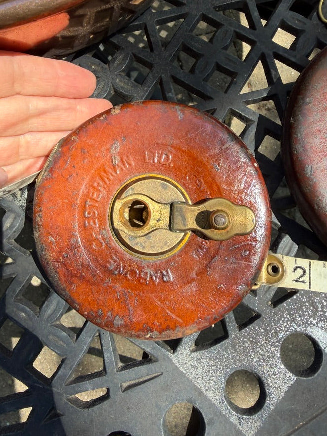 A close-up of a vintage red metal measuring tape reel held by a hand, with a measuring tape extending out. The reel has embossed text reading Chesterman, LTD