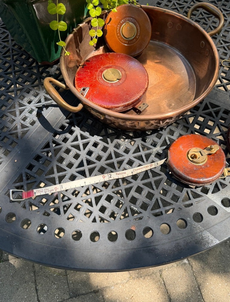 A vintage copper pot with two round red-and-gold metal lids resting inside it, placed on a black metal patio table outdoors. A measuring tape is also visible on the table.